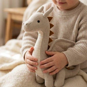 May include: A plush dragon toy in shades of gray and beige. The dragon has a long neck, wings, and brown accents on its back. The toy is held by a person wearing a beige sweater.