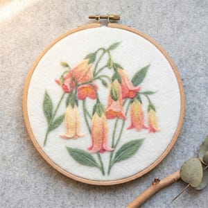 May include: A needle felted wall hanging in a circular wooden embroidery hoop featuring a cluster of bell-shaped flowers in shades of soft pink, peach, and yellow with sage green stems and leaves on a white felt background.
