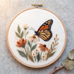 Puede incluir: Un bastidor de bordado redondo de madera que contiene tela blanca con una mariposa monarca de lana fieltrada en naranja, negro y blanco. La mariposa descansa sobre un grupo de flores de lana fieltrada en tonos naranja, crema y hojas verde salvia.