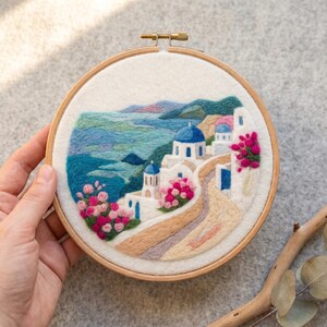 Può includere: Un telaio da ricamo circolare contenente un paesaggio in feltro ad ago raffigurante edifici bianchi con cupole blu che si affacciano su un mare blu. La scena presenta fiori rosa e rossi vivaci, un sentiero beige tortuoso e colline verdi.