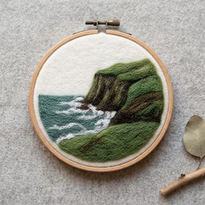 May include: A needle felted landscape art piece in a circular wooden embroidery hoop. The scene depicts a green cliffside meeting a teal ocean with white foam waves crashing against the dark brown rocky base, all set on a white felt background.
