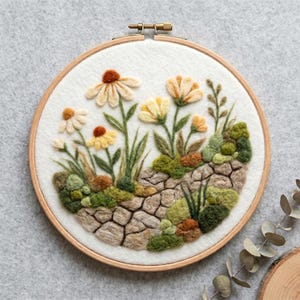 May include: A circular wooden embroidery hoop containing a needle-felted scene of white and yellow flowers growing from a textured stone path. The design features green mossy patches, brown accents, and tall stems on a white felt background.