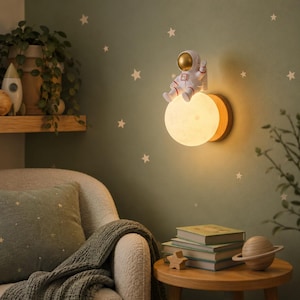 May include: A decorative wall lamp with a white astronaut figure seated on a glowing, moon-shaped light. The lamp has a wooden backing and is mounted on a green wall with white star stickers. The astronaut has a gold helmet and red detailing.