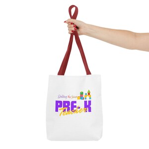 May include: A white tote bag with dark red handles featuring a graphic of colorful building blocks and the text Building the future PRE-K Teacher in purple and yellow lettering. The design is centered on the front of the white fabric bag.