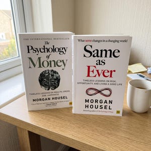 May include: Two hardcover books, "The Psychology of Money" and "Same as Ever," by Morgan Housel, are displayed on a wooden surface. The first book features a brain graphic, while the second has an infinity symbol. A white mug with a dark liquid sits nearby.