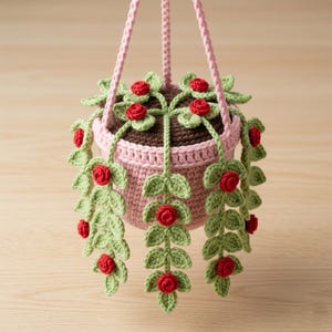 May include: A crocheted hanging plant in a light pink pot with a dark brown top. The plant features trailing green vines with small, vibrant red rose blossoms. The entire structure is suspended by three light pink crocheted cords attached to the rim.