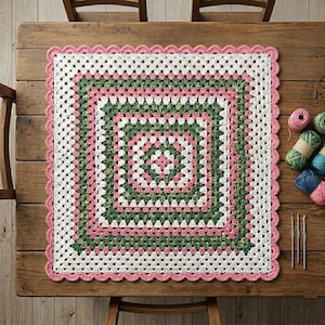 May include: A square crochet blanket featuring a granny square pattern in alternating bands of white, sage green, and soft pink. The blanket is finished with a scalloped pink border. Three crochet hooks and several yarn balls are nearby.