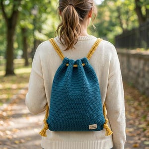May include: A teal crochet drawstring backpack featuring a textured stitch pattern and thick mustard yellow rope straps. The bag includes a small rectangular label at the bottom right corner and is worn over a cream-colored knit sweater.