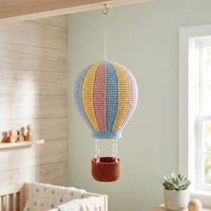 May include: A crocheted hot air balloon hanging from a gold hook. The balloon features vertical stripes in alternating pastel blue, yellow, and pink yarn. A small, solid brown crocheted basket is suspended from the balloon by four white strings.