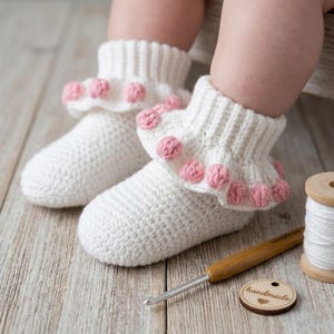 May include: A pair of white crocheted booties featuring a ribbed cuff and a ruffled edge adorned with small pink pom-poms. The booties are crafted with a textured stitch pattern and are displayed alongside a wooden crochet hook and white yarn.