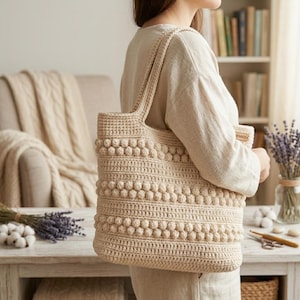 May include: A beige crochet tote bag featuring a textured pattern of horizontal rows of bobble stitches alternating with flat double crochet stitches. The bag has two integrated shoulder straps and a small rectangular tag on the side.