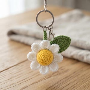 May include: A crochet daisy keychain featuring a bright yellow center, white petals, and a single green leaf. The flower is attached to a silver-toned metal split ring and a lobster clasp, providing a secure way to attach the item to keys or bags.