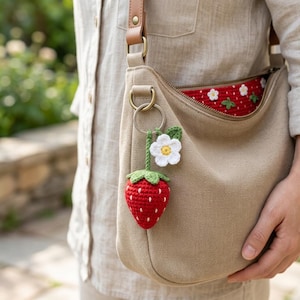 May include: A beige canvas crossbody bag with a zipper closure and a red interior lining featuring a white strawberry pattern. A crocheted strawberry keychain with a white flower and green leaves is attached to the bag's gold metal D-ring.