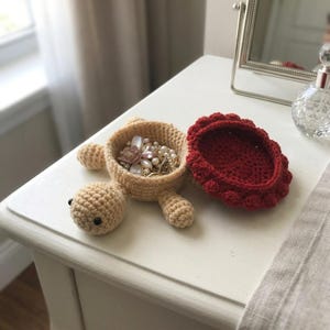 Può includere: Un portagioie all'uncinetto a forma di tartaruga color beige con un coperchio rimovibile a forma di guscio rosso scuro. Il contenitore aperto presenta un motivo a punti testurizzato e contiene vari gioielli con dettagli floreali e perle.