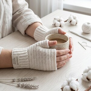 May include: A pair of off-white fingerless gloves made of crocheted yarn with a ribbed cuff. A small rectangular fabric label is attached to the wrist, featuring the printed text "HANDMADE BEGINNER GLOVES" in a clear, dark sans-serif font.