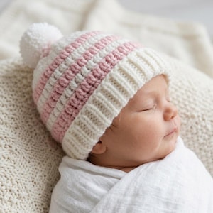 May include: A crocheted beanie hat featuring horizontal stripes in soft pink and cream white. The hat has a ribbed brim and a large, fluffy pom-pom attached to the top. The texture is consistent throughout, showcasing a classic yarn knit pattern.