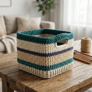 May include: A square, hand-crocheted storage basket featuring horizontal stripes in cream, dark teal, and navy blue. The basket is constructed with a thick, textured yarn and includes two integrated side handles for carrying and organizing items.
