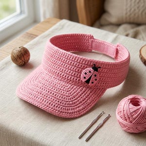 May include: A pink crocheted visor featuring a small ladybug appliqué on the front panel. The visor has a structured brim and an adjustable strap with a hook-and-loop closure at the back, crafted from a consistent pink yarn throughout the design.