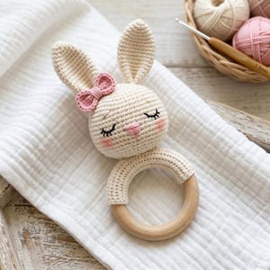 May include: A crochet baby rattle featuring a cream-colored bunny head with long ears, a small pink bow, and embroidered closed eyes. The bunny head is attached to a smooth, round, light-colored wooden ring, creating a simple, tactile toy.