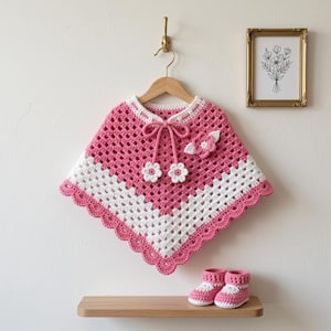 May include: A pink and white crocheted poncho featuring a V-shaped hem, a scalloped border, and a decorative tie with two small flower accents at the ends. A matching three-dimensional flower is attached to the chest. A pair of matching booties.