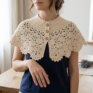 May include: A beige crochet collar featuring an intricate fan-like pattern and a scalloped edge. The garment is fastened at the front with three small, round, light brown wooden buttons, creating a structured neckline over a dark blue shirt.