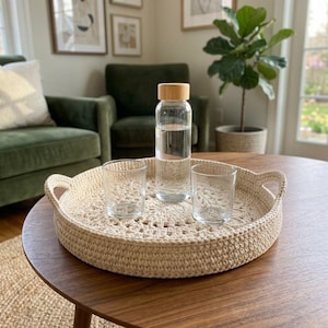 May include: A round, cream-coloured crocheted tray with two side handles, holding a clear glass water bottle with a light wood cap and two matching clear glass tumblers. The tray features a detailed, circular, open-work stitch pattern.