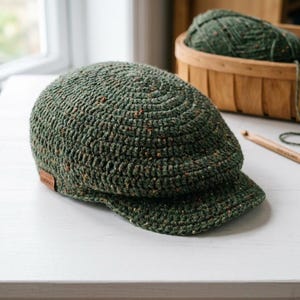 May include: A dark green crocheted flat cap featuring a textured, speckled yarn finish with small flecks of brown and orange. The cap has a rounded crown, a short stiffened brim, and a small rectangular brown leather tag on the side.