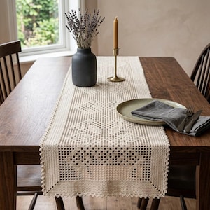 May include: A cream-colored rectangular crochet table runner with a geometric pattern, featuring a matte black vase filled with dried lavender, a tall gold-toned candle holder with a taper candle, a ceramic plate, and a grey linen napkin.