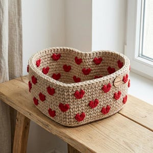 Puede incluir: Una cesta de ganchillo en forma de corazón hecha de hilo beige, con un patrón repetitivo de pequeños corazones rojos en relieve en las paredes interiores y exteriores. Una pequeña etiqueta redonda de madera clara está unida al borde superior.