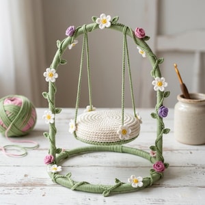 Può includere: Un set altalena decorativo all'uncinetto con una base circolare verde e un telaio ad arco avvolto in filo verde. Il telaio è decorato con piccoli fiori all'uncinetto bianchi e viola e foglie verdi. Un sedile rotondo all'uncinetto color crema pende dall'arco.