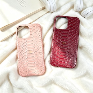 May include: Two rectangular phone cases with a textured snakeskin pattern. One case is light pink with the gold embossed letters ZH, and the other is deep red with the gold embossed letters BY. Both feature a large cutout for a camera lens.