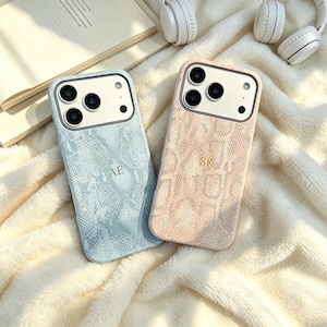 Pode incluir: Duas capas de smartphone com um padrão texturizado de pele de cobra. A capa da esquerda é azul-clara com as letras AE, e a da direita é cor de pêssego claro com as letras SK. Ambas possuem uma área branca para a câmera com três lentes.