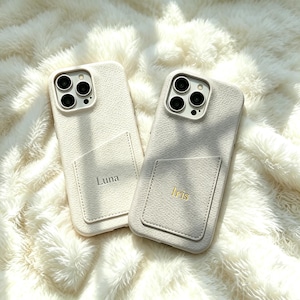 May include: Two off-white textured phone cases with integrated card slots on the back. The left case features the name Luna in grey text, while the right case features the name Iris in gold text. Both cases have cutouts for camera lenses.