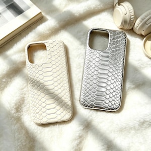 Puede incluir: Dos fundas rectangulares para smartphone con un patrón de piel de serpiente texturizado en relieve. Una funda es de color blanco roto mate y la otra es de color plata metálico. Ambas tienen un recorte cuadrado en la parte superior.