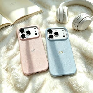 Puede incluir: Dos fundas de teléfono de cuero texturizado, una en rosa claro con las iniciales GH y otra en azul claro con las iniciales ZN. Ambas fundas cuentan con un recorte para la cámara con tres lentes circulares y un flash integrado.
