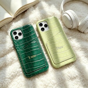 Puede incluir: Dos fundas para smartphone con textura de cocodrilo y bolsillo integrado para tarjetas. La funda izquierda es verde oscuro con el nombre Lune en dorado, y la derecha es verde claro con el nombre Thea en dorado. Ambas tienen triple recorte.