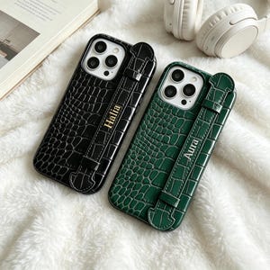 Puede incluir: Dos fundas para smartphone con textura de piel de cocodrilo brillante, una negra y otra verde oscuro. Cada funda cuenta con una correa vertical con los nombres dorados Halia y Aura impresos, junto con un recorte para la triple cámara.