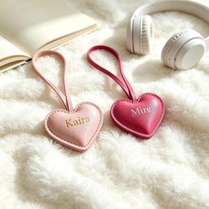 May include: Two heart-shaped leather luggage tags with loops. One tag is light pink with the name Kaira embossed in gold, and the other is dark pink with the name Mire embossed in gold. Both tags feature stitched edges and a smooth finish.