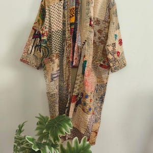 Puede incluir: Una bata estilo kimono de manga larga confeccionada con un patchwork de diversos tejidos en tonos neutros, beige y crema. La prenda presenta una compleja mezcla de patrones florales, geométricos y abstractos con costuras en rojo, azul y negro.