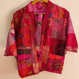 Può includere: Una giacca patchwork caratterizzata da un mix vivace di pannelli in tessuto rosa, rosso e magenta con impunture kantha bianche. Il capo presenta maniche a tre quarti, un'apertura frontale e due tasche quadrate sulla parte inferiore.