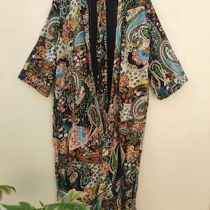 Puede incluir: Una bata estilo kimono de manga larga con un vibrante estampado floral y de cachemira multicolor sobre un fondo negro. La prenda tiene la parte delantera abierta, un dobladillo recto y costuras blancas visibles en la tela.