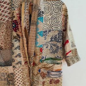 May include: A long-sleeved jacket constructed from various patches of fabric featuring diverse patterns including spirals, floral motifs, and geometric shapes in shades of beige, cream, blue, red, and black, finished with visible white running stitch quilting.