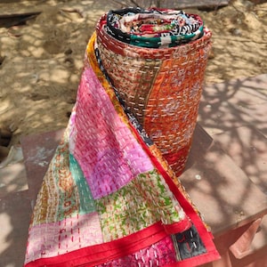 Op de afbeelding: Een opgerolde patchwork quilt met een verscheidenheid aan kleurrijke stoffen panelen in tinten roze, oranje, groen en rood. Het textiel is samengevoegd met zichtbare witte stiksteken, wat zorgt voor een getextureerd, doorgestikt uiterlijk.