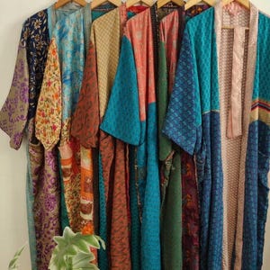 May include: A collection of six long, open-front robes hanging on wooden hangers. Each robe features a unique patchwork design with various colorful patterns, including floral, geometric, and paisley prints in shades of blue, pink, orange, and gold.