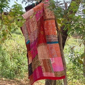 Op de afbeelding: Een rechthoekig textiel met een patchworkontwerp van verschillende stoffen vierkanten in tinten rood, oranje, roze en blauwgroen. De stof vertoont ingewikkelde patronen en zichtbare details van rijgsteken over het gehele oppervlak.