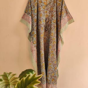 Könnte beinhalten: Ein bodenlanges Kaftan-Kleid mit hellblauer Grundfarbe und einem sich wiederholenden Blumenmuster in Gelb- und Brauntönen. Das Kleidungsstück hat weite Ärmel, einen V-Ausschnitt und ein Taillenband mit Kordelzug und Quasten sowie einen gestreiften Saum.