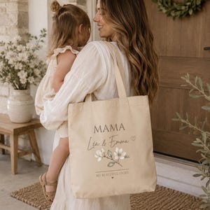 May include: A beige canvas tote bag featuring the printed text MAMA, the names L&eacute;a & Emma, a small heart icon, a floral illustration of white blossoms with green leaves, and the text MY BEAUTIFUL STORY with a tiny heart underneath.