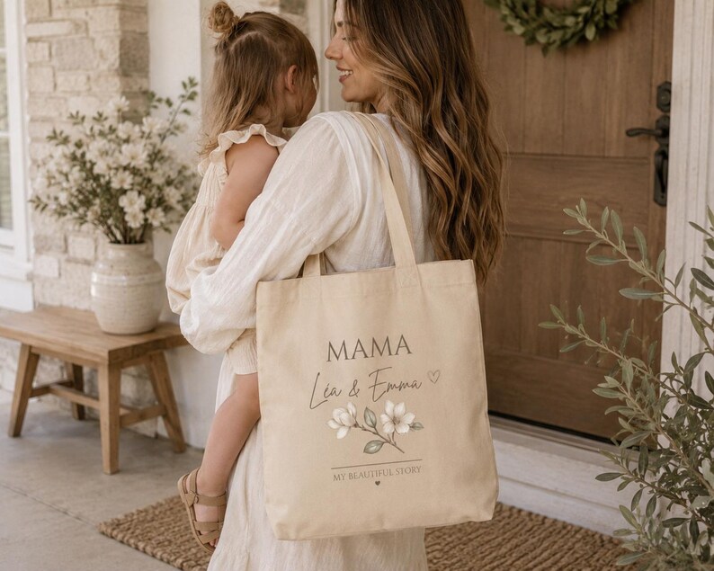 May include: A beige canvas tote bag featuring the printed text MAMA, the names L&eacute;a & Emma, a small heart icon, a floral illustration of white blossoms with green leaves, and the text MY BEAUTIFUL STORY with a tiny heart underneath.