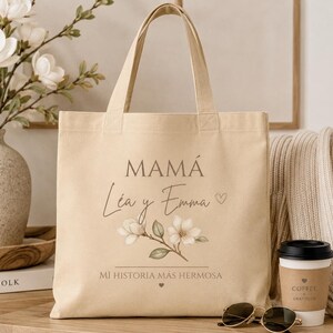 May include: A beige canvas tote bag featuring the text MAM&Aacute;, L&eacute;a y Emma, and MI HISTORIA M&Aacute;S HERMOSA in dark brown lettering. The design includes a small heart icon and a botanical illustration of white magnolia flowers with green leaves.
