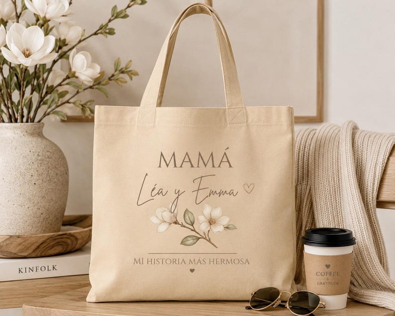 May include: A beige canvas tote bag featuring the text MAM&Aacute;, L&eacute;a y Emma, and MI HISTORIA M&Aacute;S HERMOSA in dark brown lettering. The design includes a small heart icon and a botanical illustration of white magnolia flowers with green leaves.